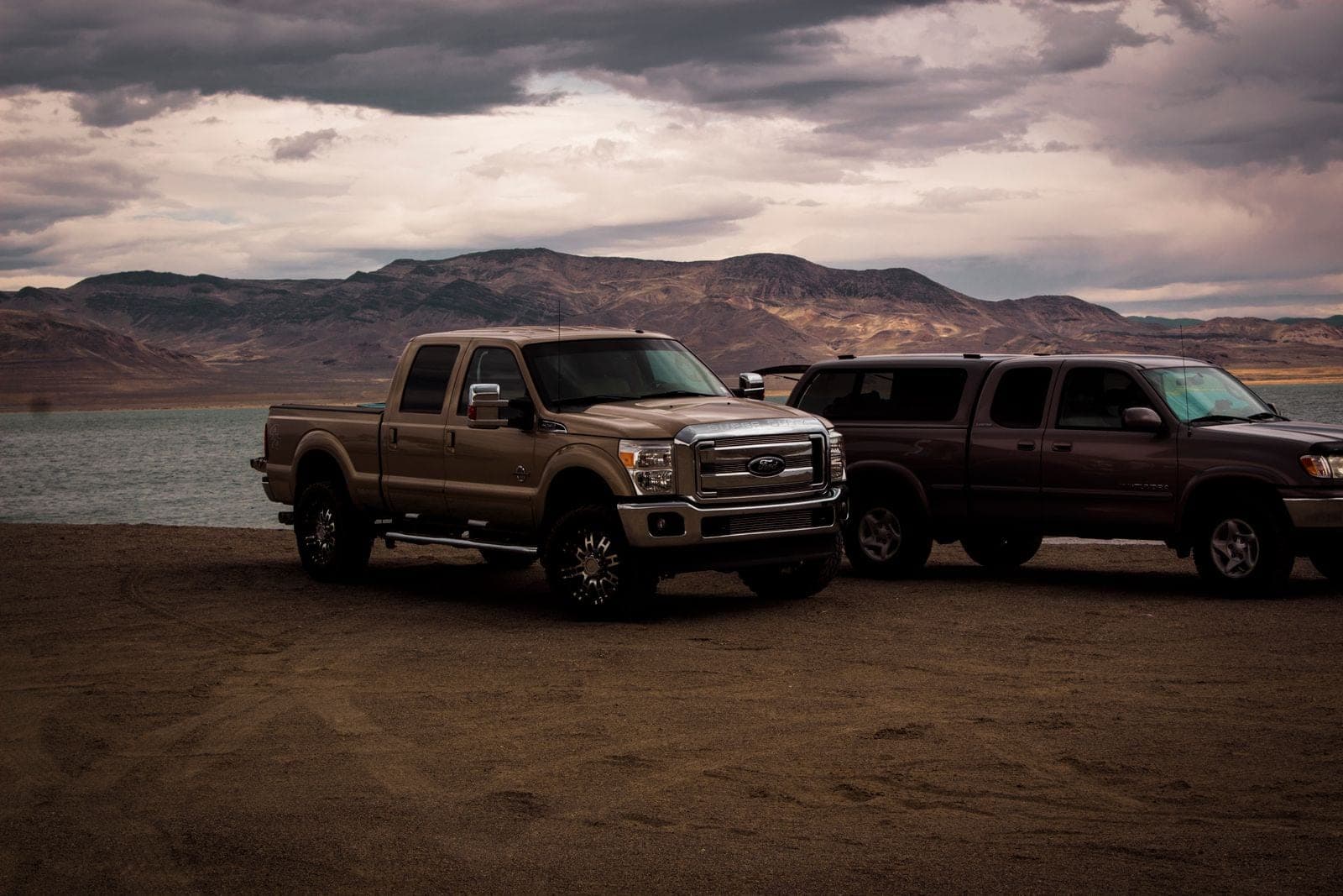 Two pickup trucks parked on a gravel shore under heavy clouds